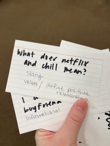 Hand holding index cards with questions. The index card on top reads "What does Netflix and Chill mean? Slang, Values/Define Positive Relationships"