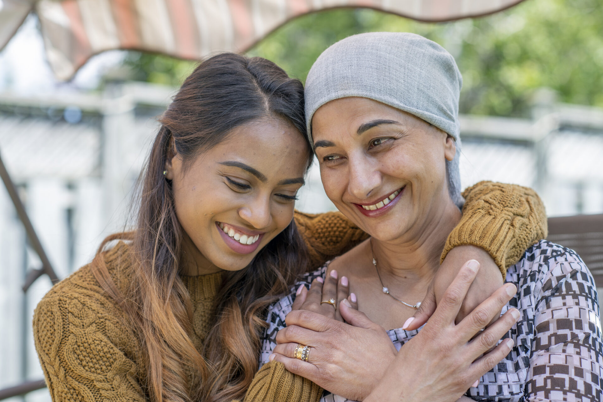 Cancer Patient Sitting Outside with Her Daughter A senior woman battling Cancer is embraced by her daughter as they sit outside enjoying the sunshine. The senior woman is dressed casually and has a head scarf on while her daughter hugs her from behind. Both are smiling as they enjoy the close moment together.